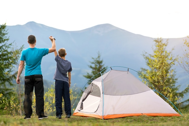 Giovane papà e suo figlio figlio in piedi vicino alla tenda da campeggio con le mani alzate mentre fanno un'escursione insieme in montagna.