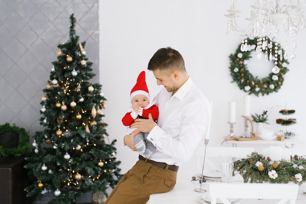 Giovane papà felice che tiene il suo piccolo figlio in costume di Santa vicino all'albero di Natale in salone