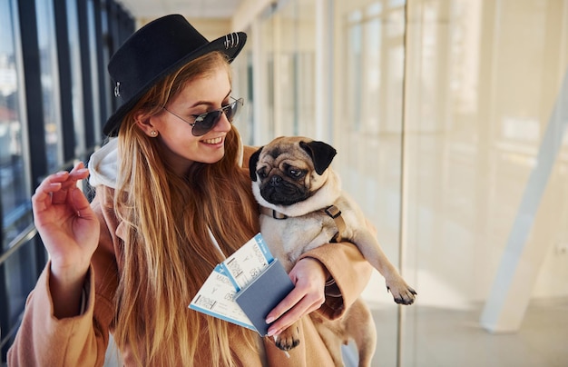Giovane passeggero femminile in abiti caldi con in mano i biglietti e un simpatico cane in mano nella hall dell'aeroporto.