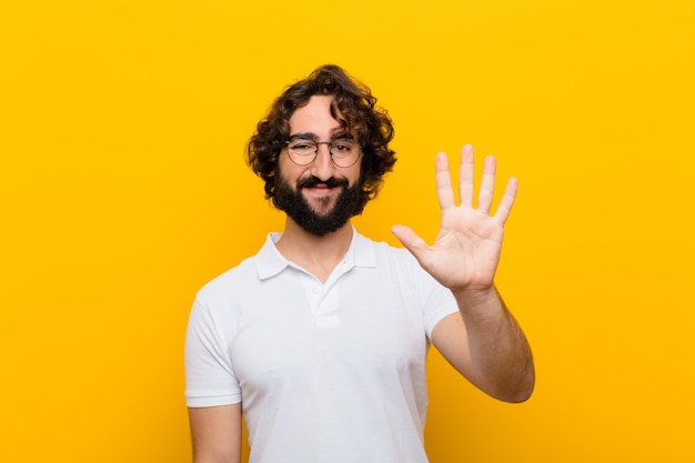 Giovane pazzo che sorride e che sembra amichevole, mostrando numero cinque o quinto con la mano in avanti, conto alla rovescia contro la parete gialla