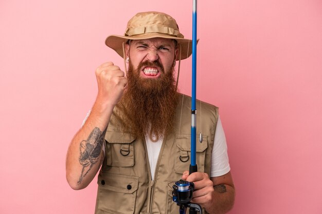 Giovane pescatore caucasico dello zenzero con la barba lunga che tiene un'asta isolata su fondo rosa che mostra pugno alla macchina fotografica, espressione facciale aggressiva.