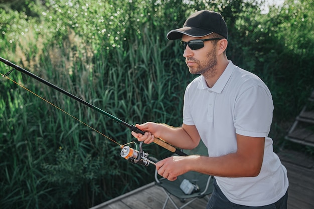 Giovane pescatore che pesca sul lago o sul fiume. Immagine del ragazzo adulto concentrato occupato che gira la bobina durante la caccia in acqua. Prepararsi a pescare un gustoso pesce prelibato.