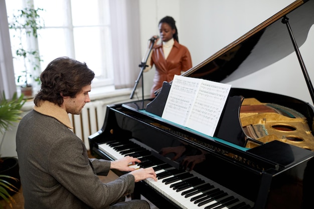 Giovane pianista seduto davanti allo spartito e che suona la musica al pianoforte con una donna africana che canta al microfono in sottofondo