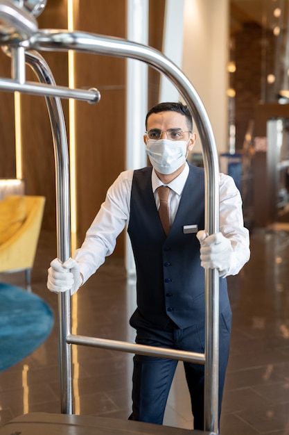 Giovane portiere in uniforme, occhiali e maschera protettiva che spinge il carrello con i bagagli mentre si muove verso le stanze degli ospiti all'interno dell'hotel