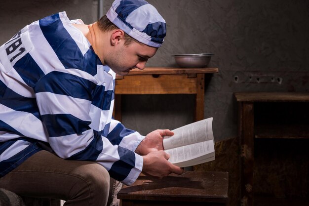 Giovane prigioniero maschio che indossa l'uniforme della prigione leggendo un libro o una Bibbia mentre è seduto su un letto vicino al comodino con piatti di alluminio in una cella di prigione