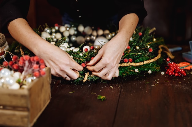 Giovane progettista sorridente sveglio della donna che prepara la corona dell'albero sempreverde di Natale, produttore delle decorazioni di Natale con le proprie mani, corona di Natale per la festa, la celebrazione del nuovo anno.