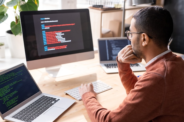 Giovane programmatore pensieroso seduto sul posto di lavoro e guardando le informazioni sullo schermo del computer durante l'analisi