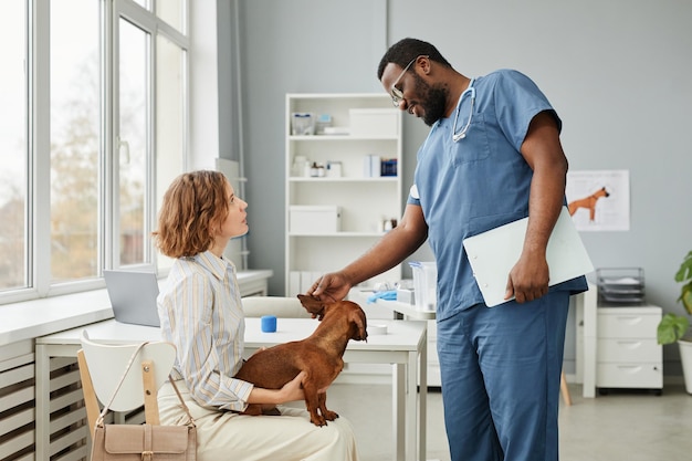 Giovane proprietario di animali domestici seduto su una sedia in cliniche veterinarie contemporanee e guardando il veterinario mentre si consulta sul suo cane