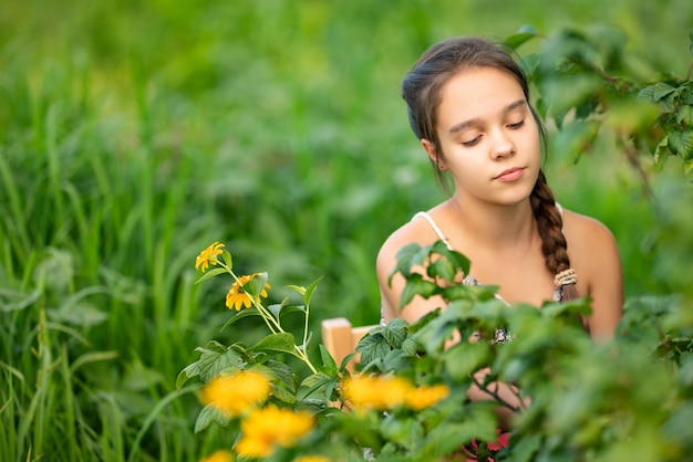 Giovane ragazza adolescente con una treccia marrone si siede in un giardino vicino a un cespuglio e fiori