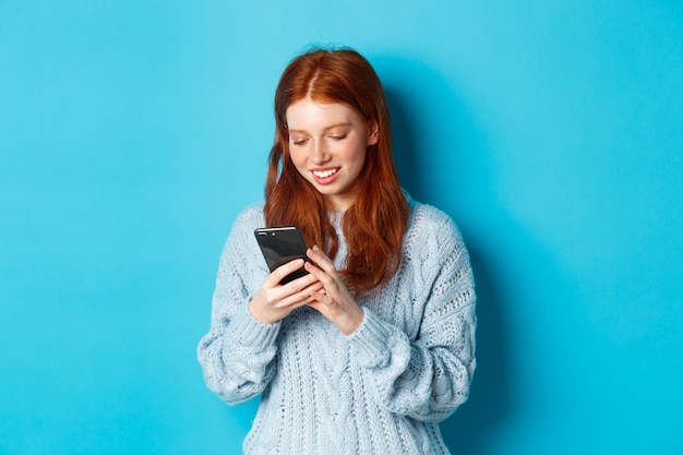 Giovane ragazza adolescente rossa leggendo il messaggio su smartphone e sorridente, utilizzando il telefono cellulare e in piedi su sfondo blu.