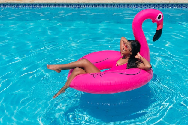 Giovane ragazza africana sexy allegra che indossa il costume da bagno fucsia divertendosi nella piscina all'aperto dell'hotel in fenicottero gonfiabile fucsia.