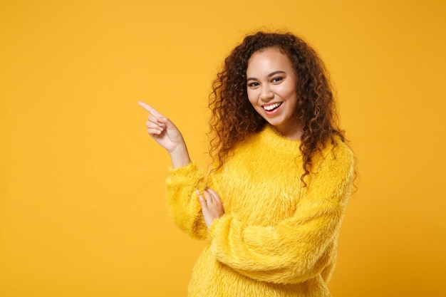 Giovane ragazza afroamericana allegra in maglione di pelliccia in posa isolata su sfondo giallo arancio in studio. Persone sincere emozioni concetto di stile di vita. Mock up spazio di copia. Puntando il dito indice da parte.