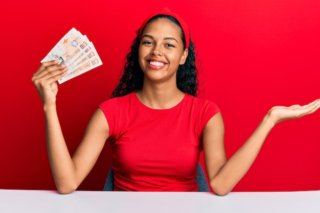 Giovane ragazza afroamericana che tiene le sterline del Regno Unito seduta sul tavolo celebrando il successo con un sorriso felice e l'espressione del vincitore con la mano alzata