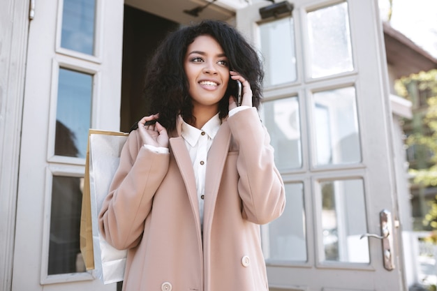 Giovane ragazza afroamericana in piedi con il telefono e pacchetti in mano Bella ragazza sorridente in cappotto che parla sul suo telefono cellulare Ritratto di bella signora con i capelli ricci scuri e il cellulare in mano