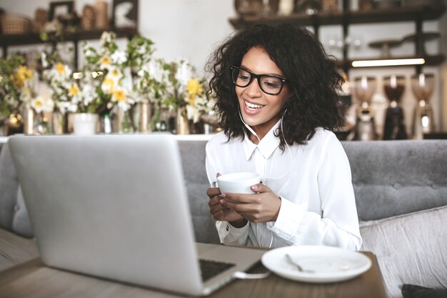 Giovane ragazza afroamericana in vetri che si siedono nel ristorante con il computer portatile e la tazza di caffè nelle mani