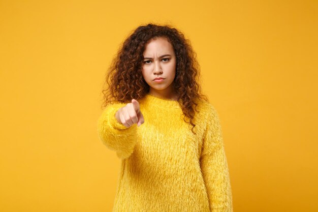 Giovane ragazza afroamericana insoddisfatta in maglione di pelliccia in posa isolata su sfondo giallo arancione parete ritratto in studio. Concetto di stile di vita delle persone. Mock up spazio di copia. Puntare il dito indice sulla fotocamera.