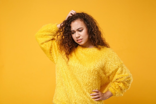 Giovane ragazza afroamericana preoccupata in maglione di pelliccia in posa isolata su sfondo giallo arancio. Persone sincere emozioni concetto di stile di vita. Mock up spazio di copia. Mettere una mano sulla testa, guardare in basso.