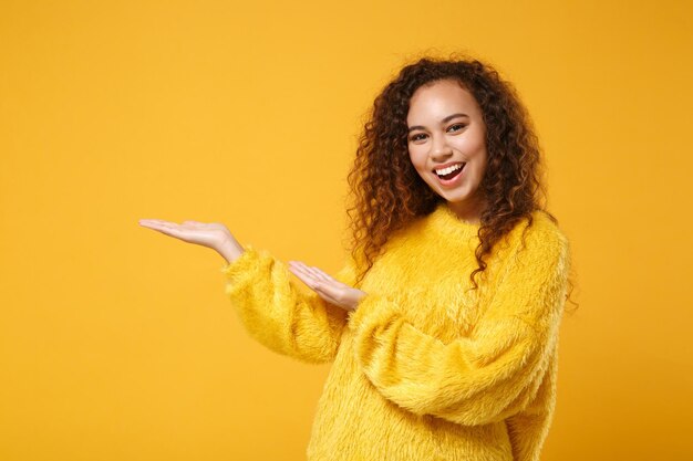 Giovane ragazza afroamericana ridente allegra in maglione di pelliccia in posa isolata su sfondo giallo arancione muro, ritratto in studio. Concetto di stile di vita delle persone. Mock up spazio di copia. Indicando le mani da parte.