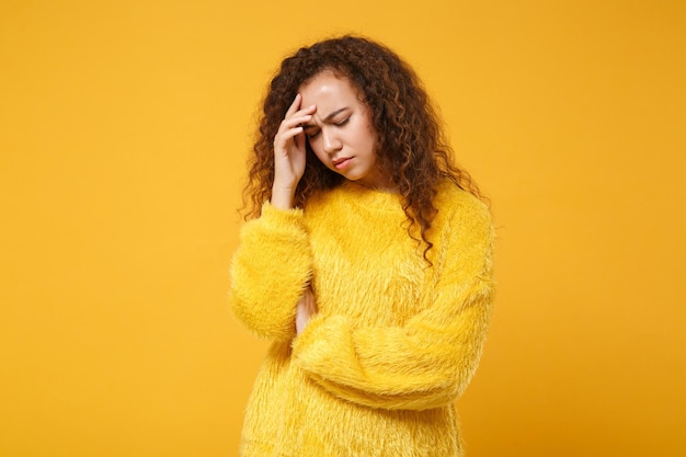 Giovane ragazza afroamericana stanca dispiaciuta in maglione di pelliccia in posa isolata su sfondo giallo arancione in studio. Concetto di stile di vita delle persone. Mock up spazio di copia. Tenendo gli occhi chiusi, metti la mano sulla testa.