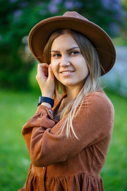 Giovane ragazza alla moda in un cappello vicino al cespuglio lussureggiante di lillà rosa in giardino. La ragazza gode dell'odore del lillà. Calda giornata estiva in giardino giovane donna con un sorriso sul viso