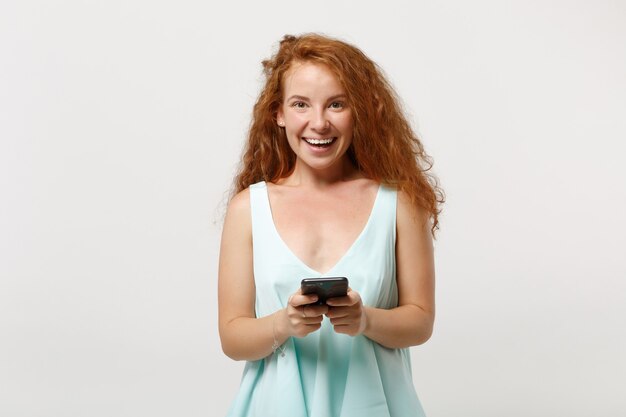 Giovane ragazza allegra della donna della testarossa in vestiti leggeri casuali che posano isolati su fondo bianco, ritratto dello studio. Concetto di stile di vita della gente. Mock up copia spazio. Utilizzando il telefono cellulare, digitando un messaggio sms.