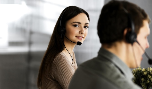 Giovane ragazza allegra in cuffia sta guardando la fotocamera, mentre è seduto alla scrivania con un collega maschio. Ritratto di operatore di call center al lavoro.