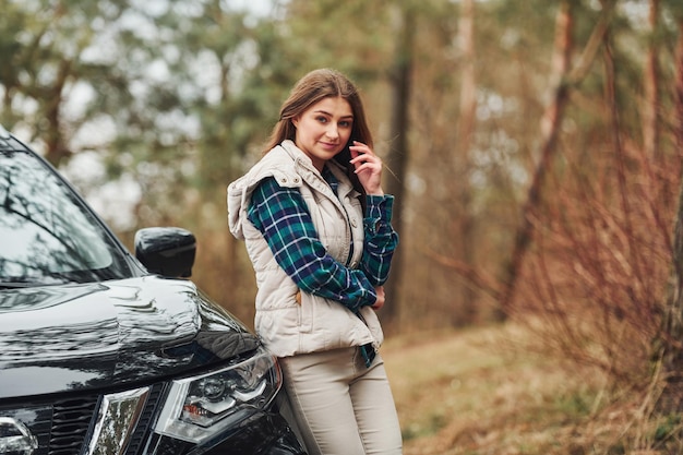 Giovane ragazza allegra in piedi vicino alla moderna auto nera all'aperto nella foresta