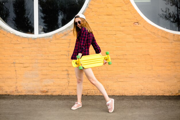Giovane ragazza allegra in posa con skateboard giallo contro il muro arancione.