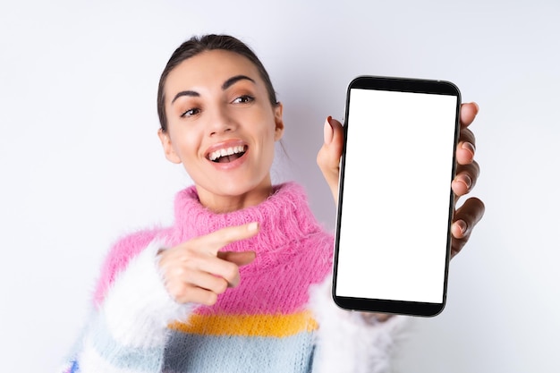 Giovane ragazza allegra in un maglione colorato brillante su sfondo bianco tiene un grande telefono a fuoco con uno schermo bianco vuoto