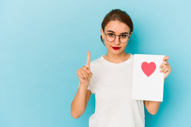 Giovane ragazza araba magra che tiene una carta di San Valentino che mostra il numero uno con il dito.