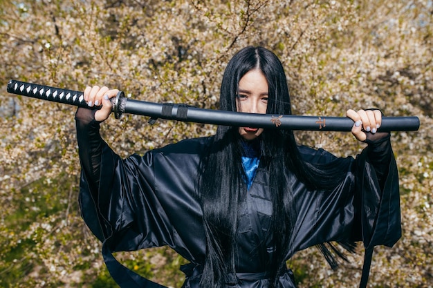 Giovane ragazza asiatica in kimono nero tradizionale con la spada sullo sfondo della natura