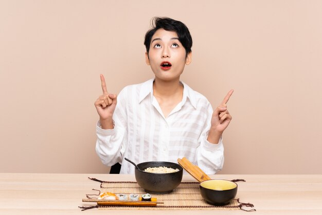 Giovane ragazza asiatica in un tavolo con una ciotola di noodles e sushi che punta con il dito indice un'ottima idea