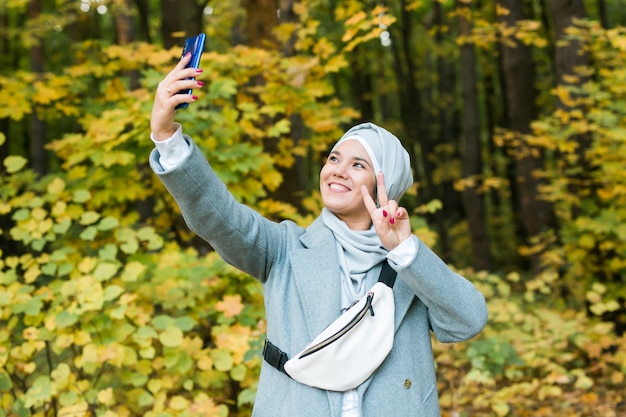 Giovane ragazza asiatica musulmana alla moda in hijab che prende un selfie sullo smartphone all'aperto nel parco autunnale