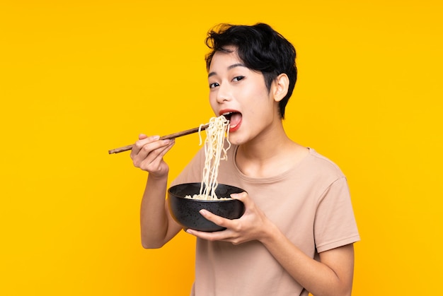 Giovane ragazza asiatica sopra la parete gialla isolata che tiene una ciotola di tagliatelle con le bacchette e che la mangia