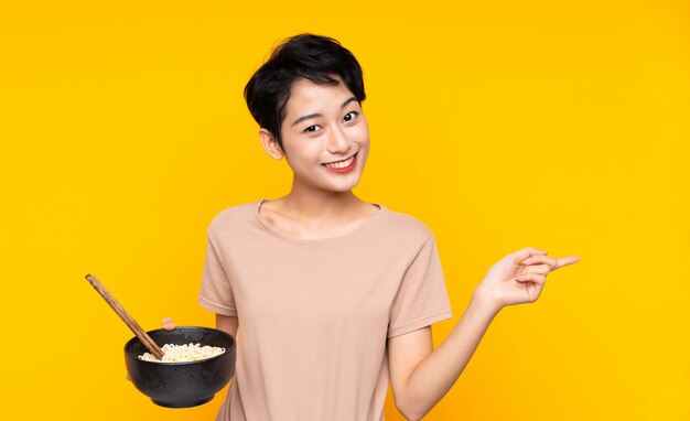 Giovane ragazza asiatica sopra la parete gialla isolata sorpresa e puntando il dito verso il lato mentre si tiene una ciotola di noodles con le bacchette