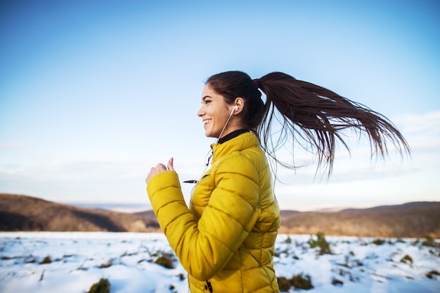 Giovane ragazza atletica attiva che pareggia in abiti sportivi di inverno sulla strada nevosa di inverno con le cuffie nella mattina soleggiata.