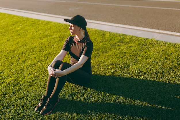 Giovane ragazza atletica in uniforme nera, berretto seduto rilassante che riposa sul prato verde nel parco del campo da golf dopo esercizi sportivi, correndo all'aperto nella soleggiata giornata estiva. Fitness, concetto di stile di vita sano.