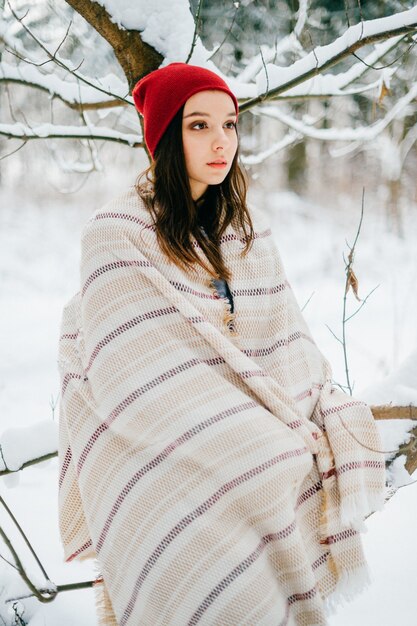 Giovane ragazza attraente che copre con un caldo mantello in posa tra i rami della neve nella foresta