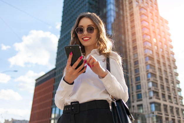 giovane ragazza attraente in occhiali da sole e camicia bianca sta utilizzando lo smartphone per strada in città