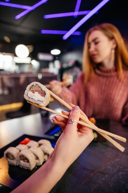 Giovane ragazza attraente in possesso di un rotolo di sushi makizushi servito su piatto nero su sfondo sushibar