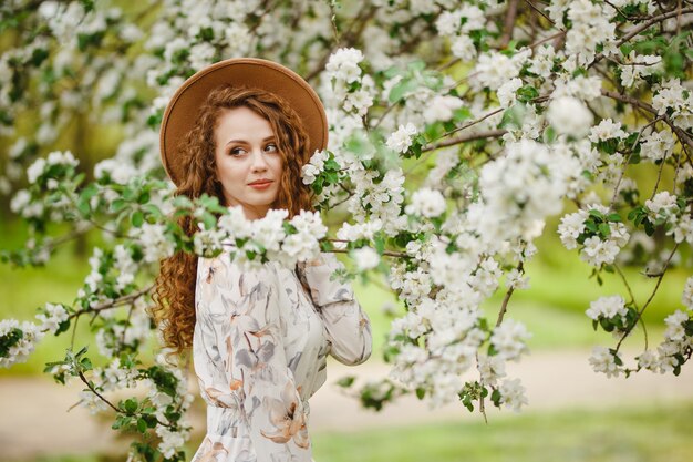 Giovane ragazza attraente si trova tra gli alberi in fiore bianchi al parco. Una donna che indossa un cappello marrone e un abito bianco gode della stagione primaverile. Giardino fresco e cambiamento del concetto di stagioni.