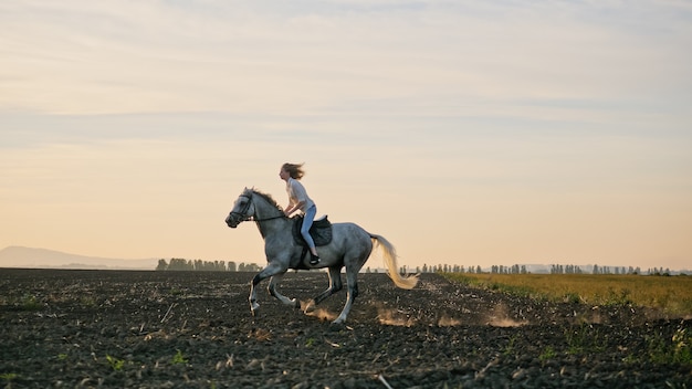 Giovane ragazza bionda a cavallo sul campo durante il tramonto. Gli zoccoli del cavallo sollevano polvere mentre corre su terreno asciutto