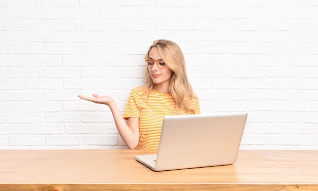 giovane ragazza bionda al computer portatile sentirsi felice e sorridente in cerca di un oggetto tenuto sulla mano sul lato