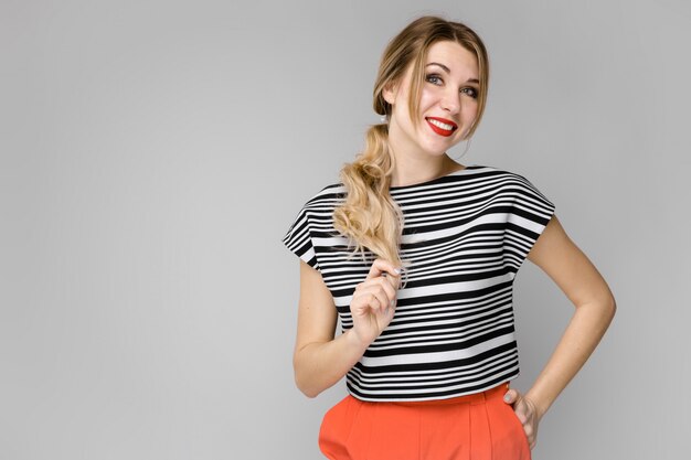Giovane ragazza bionda attraente in blusa a strisce che sorride tenendo la sua condizione dei capelli