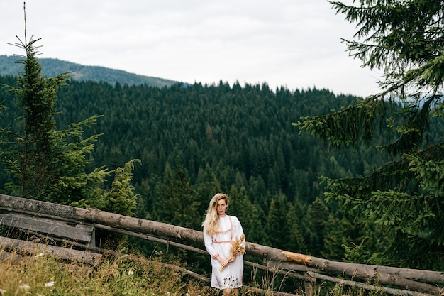 Giovane ragazza bionda attraente in vestito bianco con ricamo in posa con bouquet di spighette sul pittoresco paesaggio