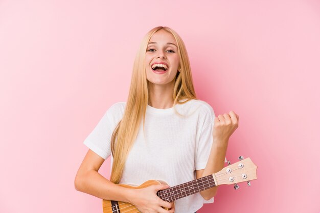 Giovane ragazza bionda che gioca ukelele che incoraggia spensierato ed eccitato. Concetto di vittoria.