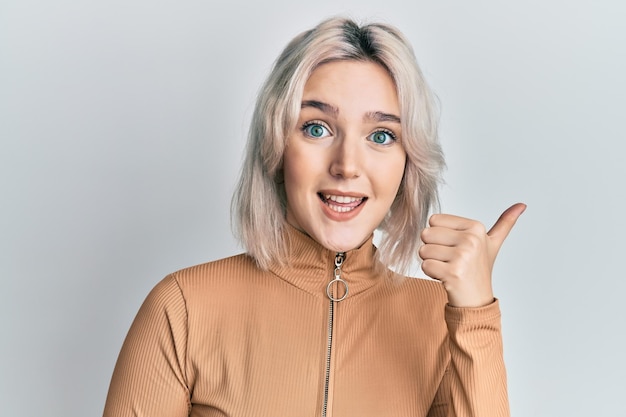 Giovane ragazza bionda che indossa abiti casual sorridente con la faccia felice che guarda e indica il lato con il pollice in su