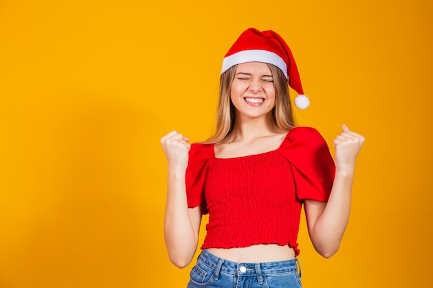 Giovane ragazza bionda che indossa il cappello di Natale celebra sorpresa e stupita per il successo con le braccia alzate. concetto di vincitore.