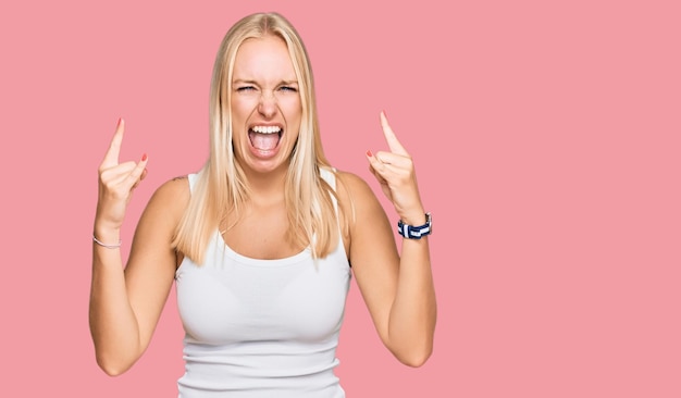 Giovane ragazza bionda che indossa uno stile casual con camicia senza maniche che grida con un'espressione pazza che fa il simbolo rock con le mani in alto. stella della musica. concetto di musica pesante.