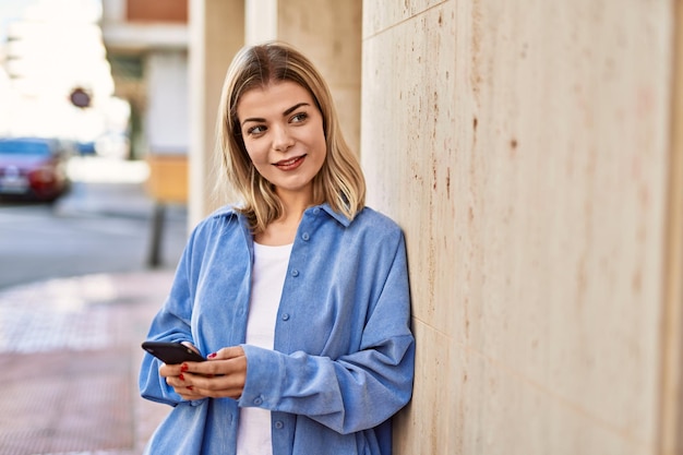 Giovane ragazza bionda che sorride felice utilizzando lo smartphone in città.